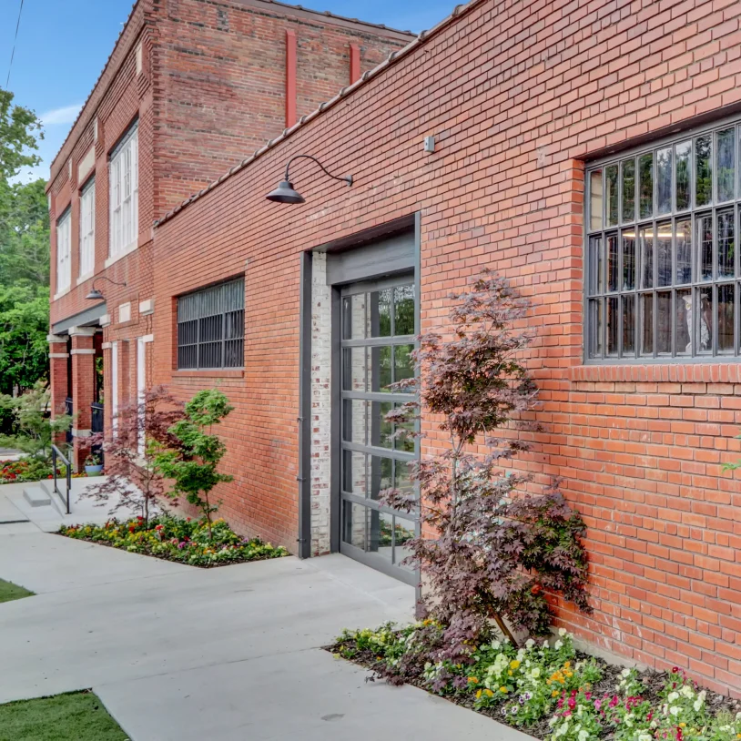 Restored brick exterior of Lofts at Creek Street with large industrial-style windows and landscaped flower beds along the sidewalk.
