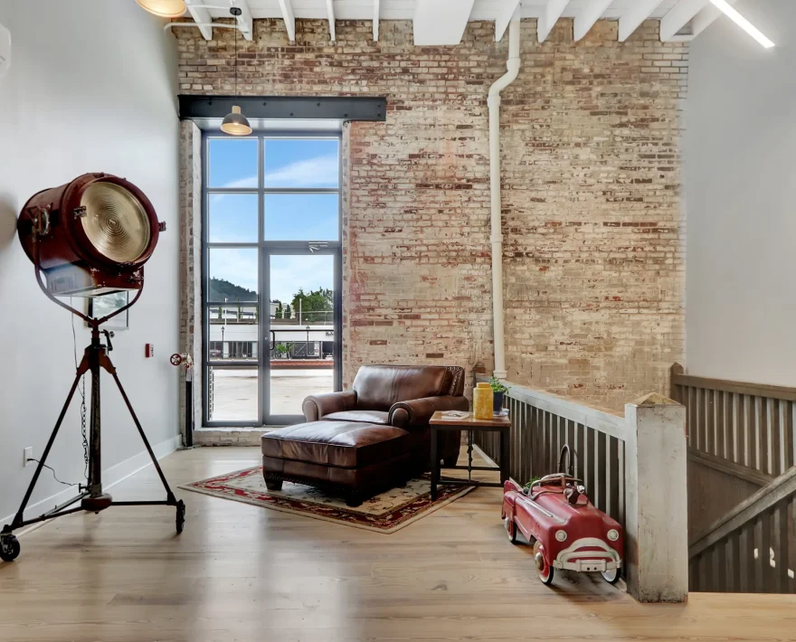 Bright upper-level lounge area with floor-to-ceiling industrial windows, exposed brick, and leather seating inside a converted historic property